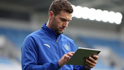 A mid-30s male staff member in a solid blue training jacket, waist-up, focused on a tablet displaying mental drills...