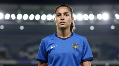 A mid-30s anonymous female player in a solid blue jersey, waist-up, focused expression, background blurred stadium...