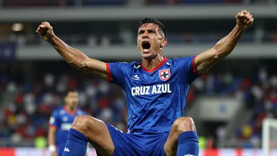 Mid-30s muscular forward, dark hair, intense eyes, wearing Cruz Azul home kit - royal blue shirt with red trim, club...