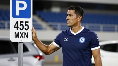 A Cruz Azul player in a navy blue jersey with white trim, mid-30s, muscular build, standing in a stadium parking lot with...