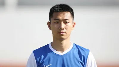 A 25-year-old coach with a calm expression, wearing a blue jersey with white sleeves and a white collar, standing in...