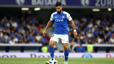 A tight waist-up shot of a young, anonymous, dark-haired, 25-30-year-old, bearded midfielder in a blue jersey with white...