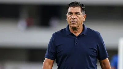 Tight waist-up shot of a stocky 40s Latin American football coach in a solid navy blue polo shirt, blurred stadium background