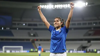 A young female soccer player in a solid blue jersey and white shorts, mid-celebration with arms raised, standing on the...
