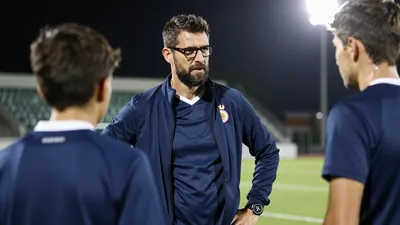 A 30-year-old, ruggedly-built, dark-haired, bearded, and bespectacled assistant coach, wearing a navy blue, long-sleeved...