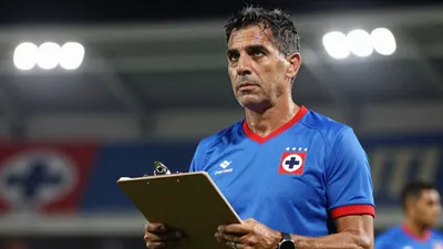 Middle-aged, fit coach with short dark hair, intense eyes, wearing Cruz Azul home kit - blue shirt with red trim, crest...