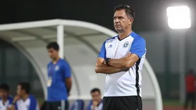 A 35-year-old, ruggedly-built, dark-haired coach with a strong jawline, wearing a white jersey with blue sleeves and a...