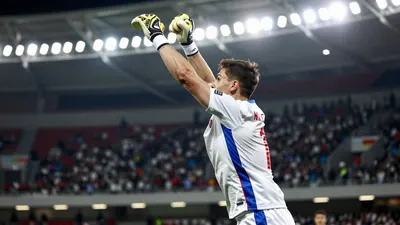 A photo of a goalkeeper in action, wearing a white jersey with a blue and red stripe, in a bright floodlit stadium, with...