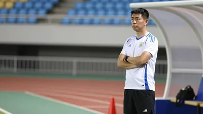 A 30-year-old male coach with a stern expression, wearing a white jersey with a blue stripe and a pair of black shorts...