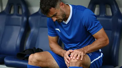 A 25-year-old male soccer player with a muscular build, wearing a solid blue jersey and shorts, sits on the bench during...