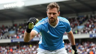A 35-year-old, 5'11" goalkeeper with a rugged build, wearing a blue jersey with white sleeves and white shorts, with a...